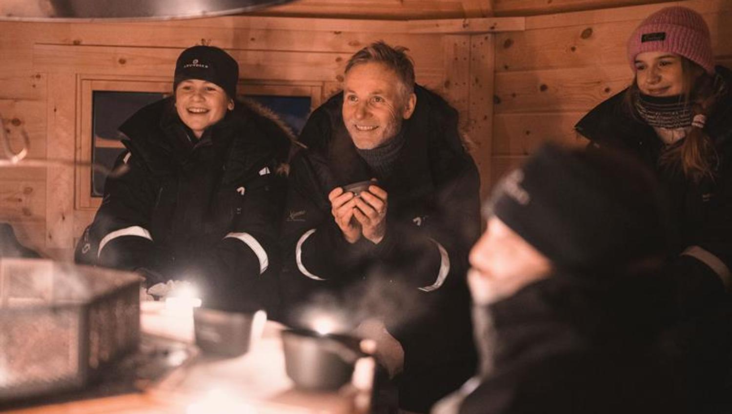 Personer sitter i en koie rundt åpen ild