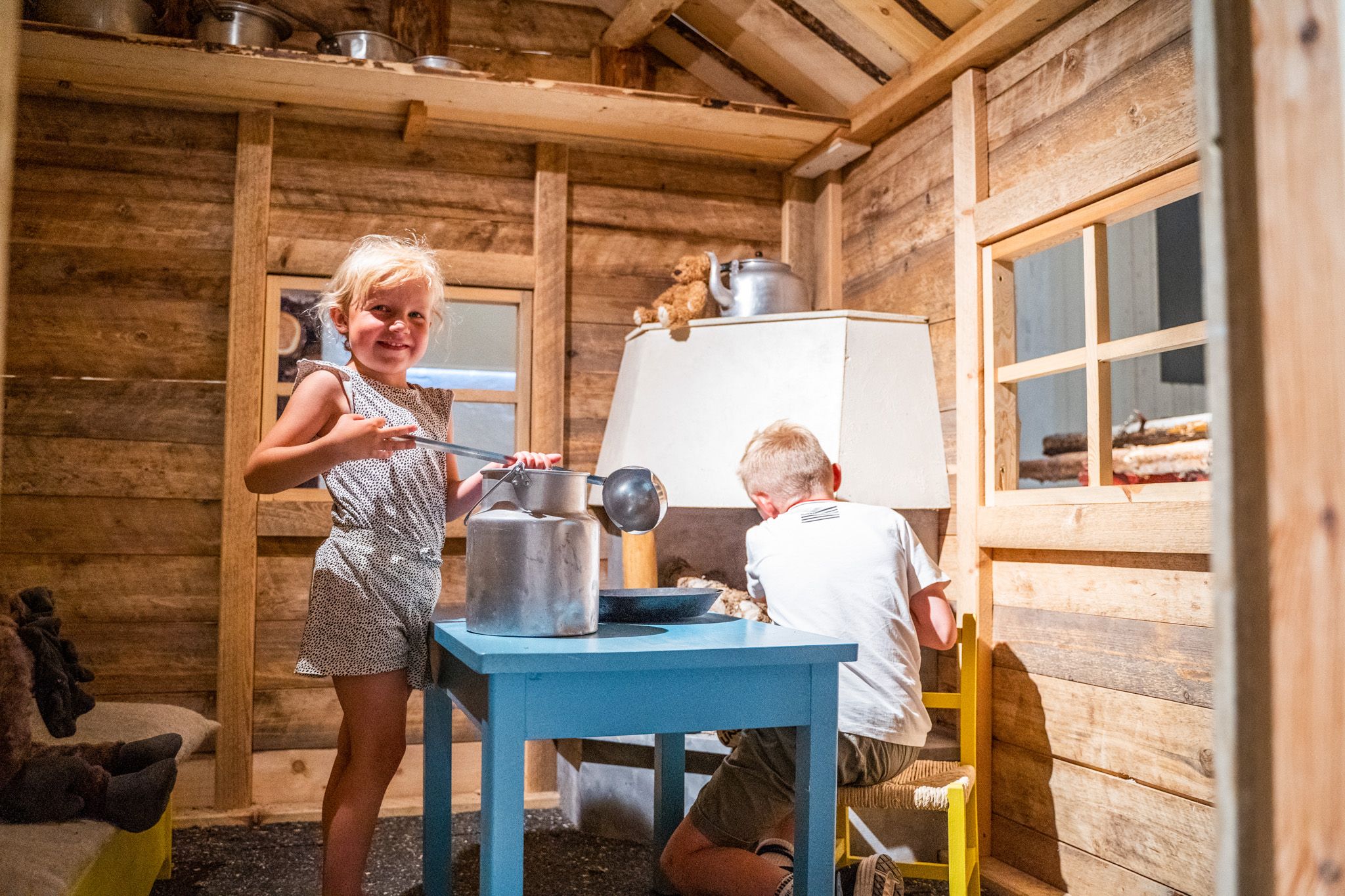 Bildet viser to barn som leker inne i et lite trehus. Jenta smiler mot kameraet mens hun holder en sleiv over en melkekanne på et blått bord. Gutten sitter ved bordet og later som han jobber med noe. Det ser ut som de leker kjøkken eller kafé.