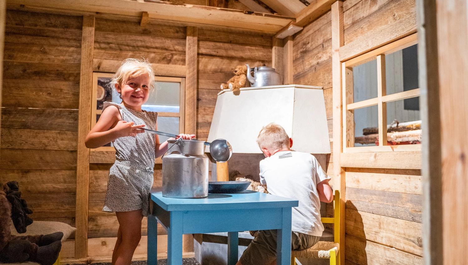Bildet viser to barn som leker inne i et lite trehus. Jenta smiler mot kameraet mens hun holder en sleiv over en melkekanne på et blått bord. Gutten sitter ved bordet og later som han jobber med noe. Det ser ut som de leker kjøkken eller kafé.