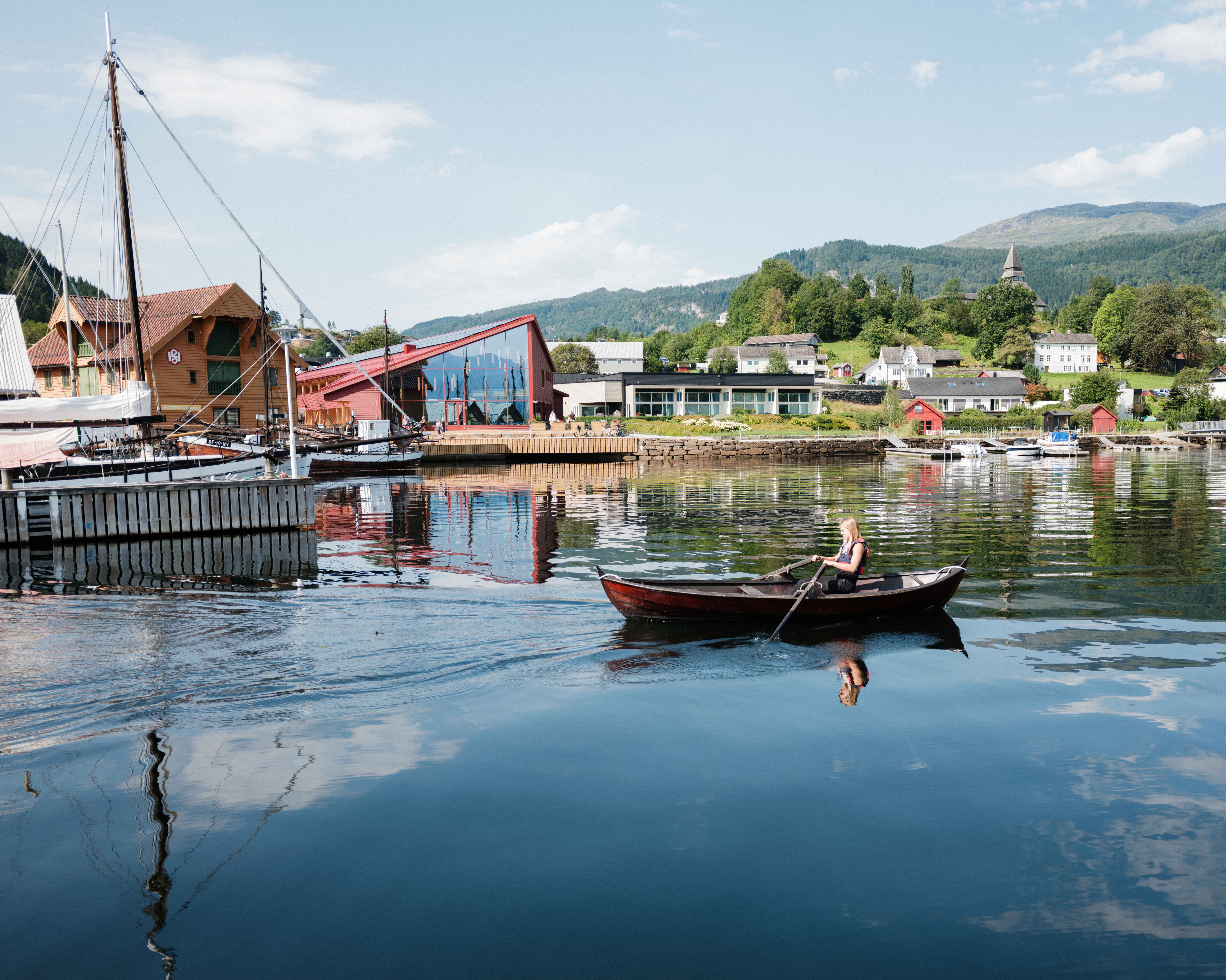 Woman rowing on the fjord with the Hardanger Maritime Centre and Norheimsund in the background – a taste of coastal culture in Hardanger.