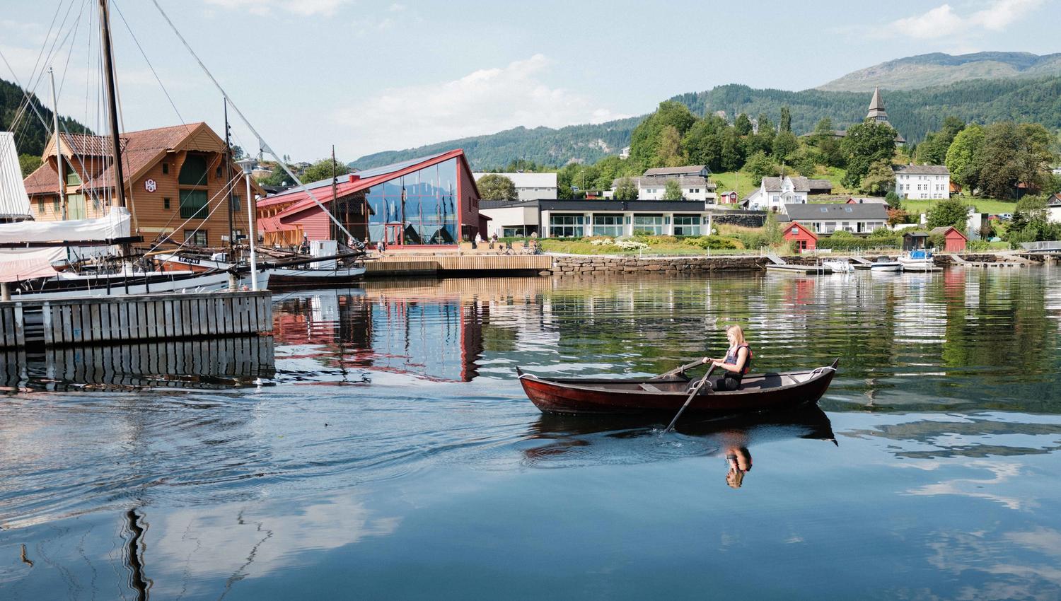Woman rowing on the fjord with the Hardanger Maritime Centre and Norheimsund in the background – a taste of coastal culture in Hardanger.