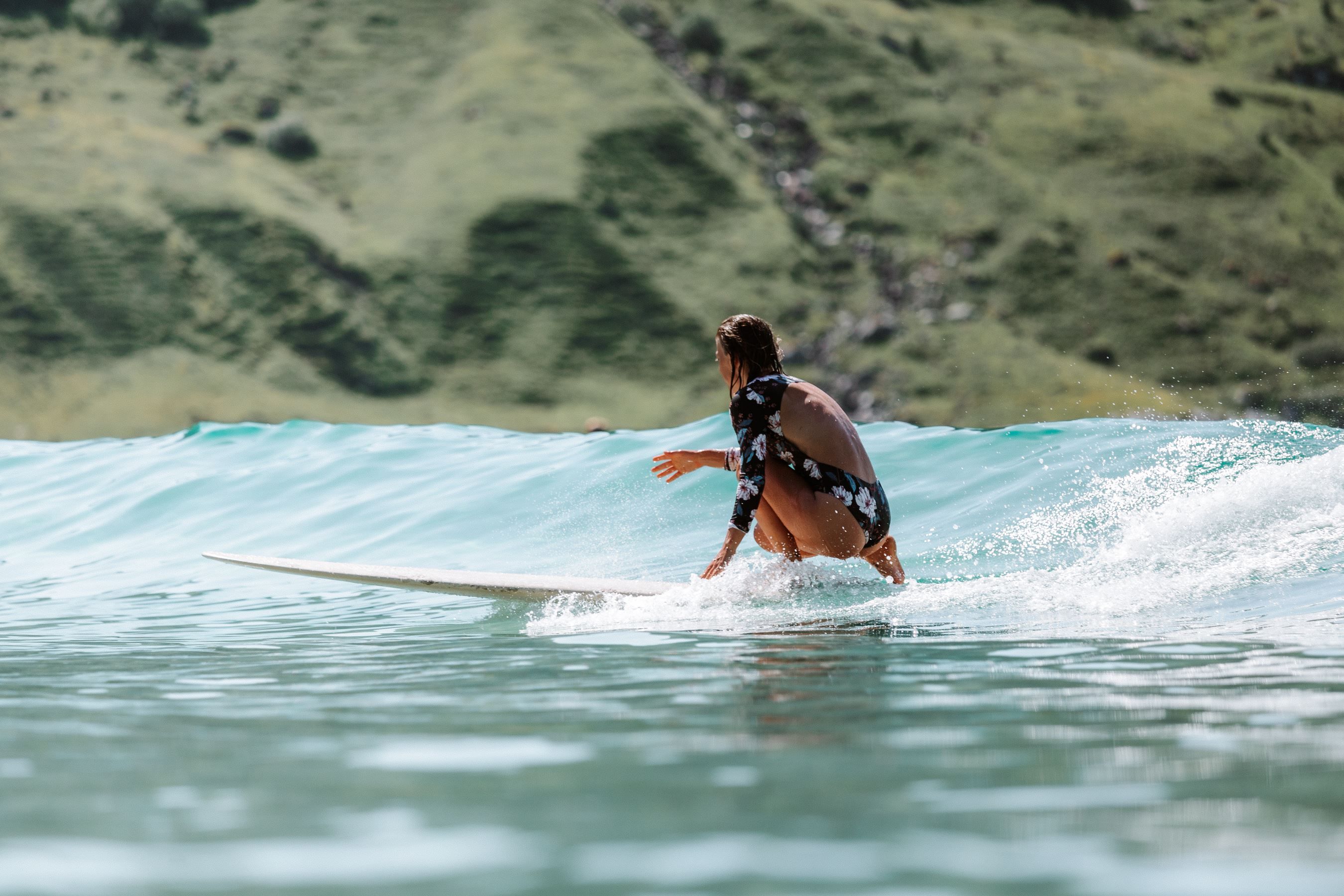 A surfer in a floral long-sleeve swimsuit crouches on a surfboard riding a small wave with green hills behind..