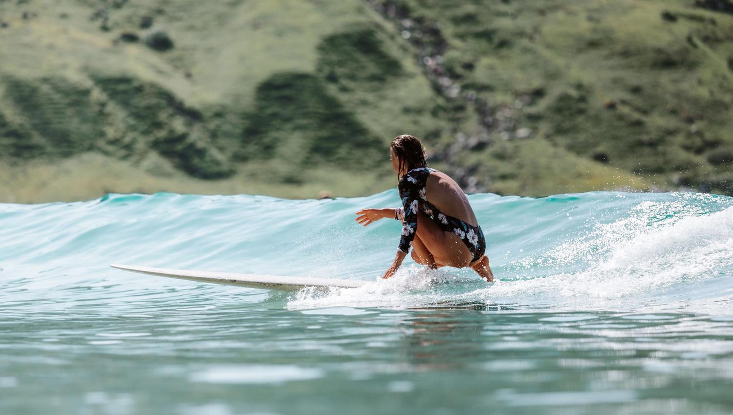 A surfer in a floral long-sleeve swimsuit crouches on a surfboard riding a small wave with green hills behind..
