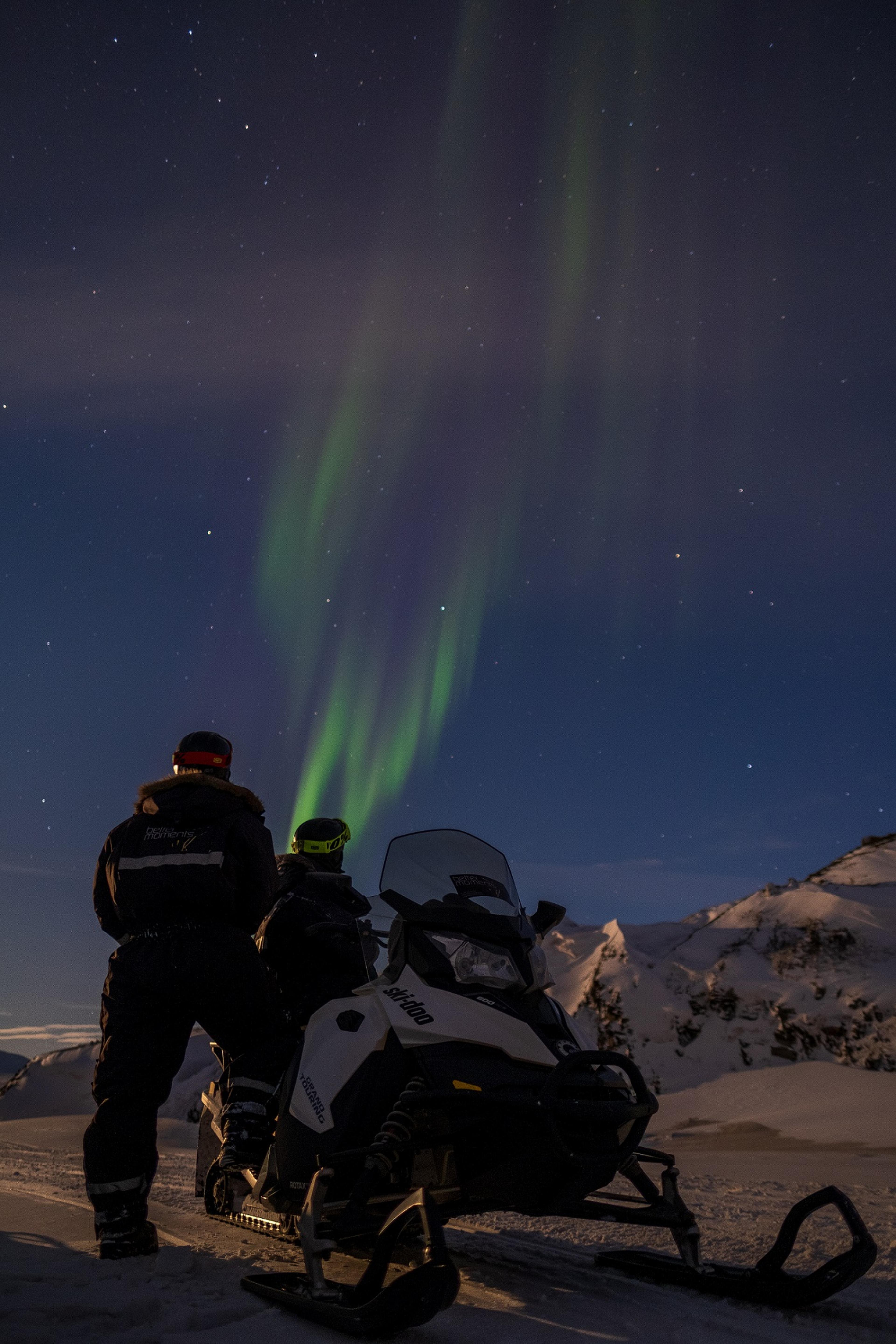 Two persons standing behind a snowmobile and looking up at northern lights in the sky