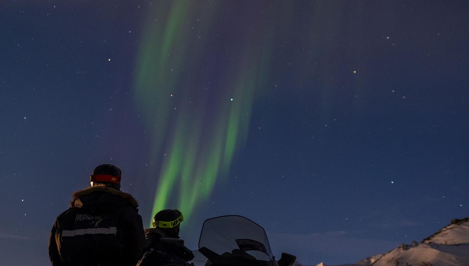 To personer som står bak en snøscooter og ser på nordlys på himmelen