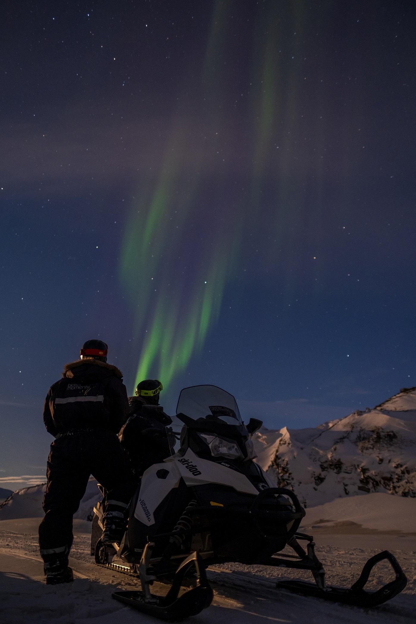 Two persons standing behind a snowmobile and looking up at northern lights in the sky