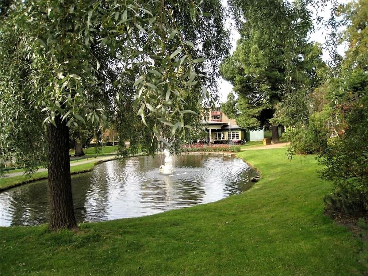 Grønn park med dam og fontene, omkranset av trær og plen, med en bygning synlig i bakgrunnen..