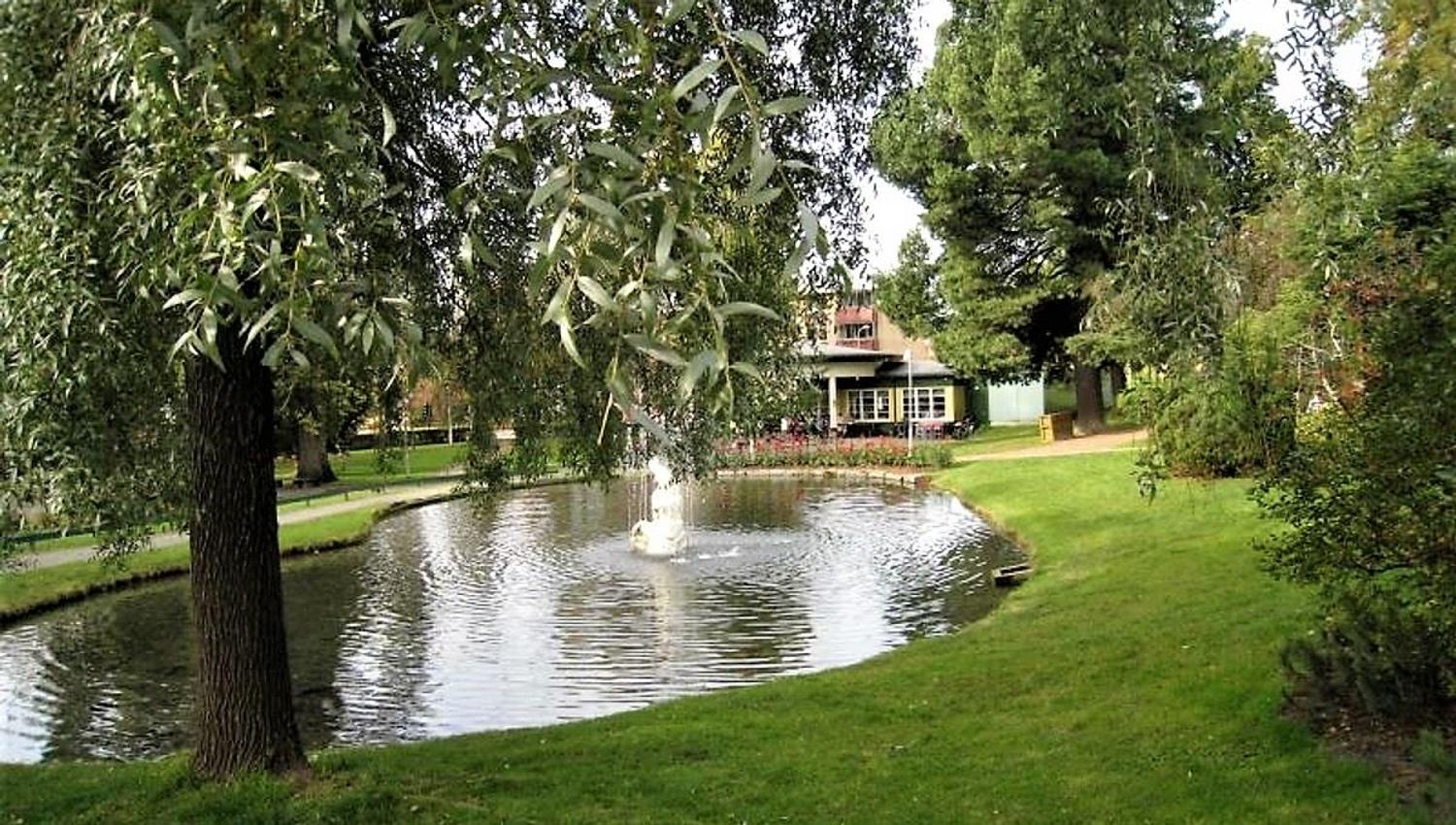 Grønn park med dam og fontene, omkranset av trær og plen, med en bygning synlig i bakgrunnen..
