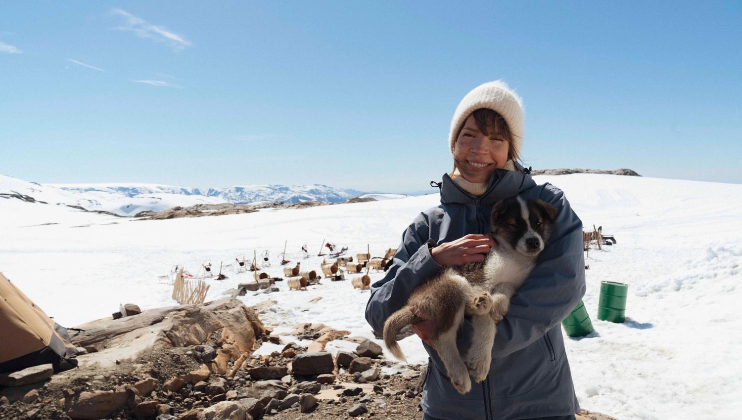 En person smiler og holder en huskyvalp med folgefonna husky camp og fjell i bakgrunnen.