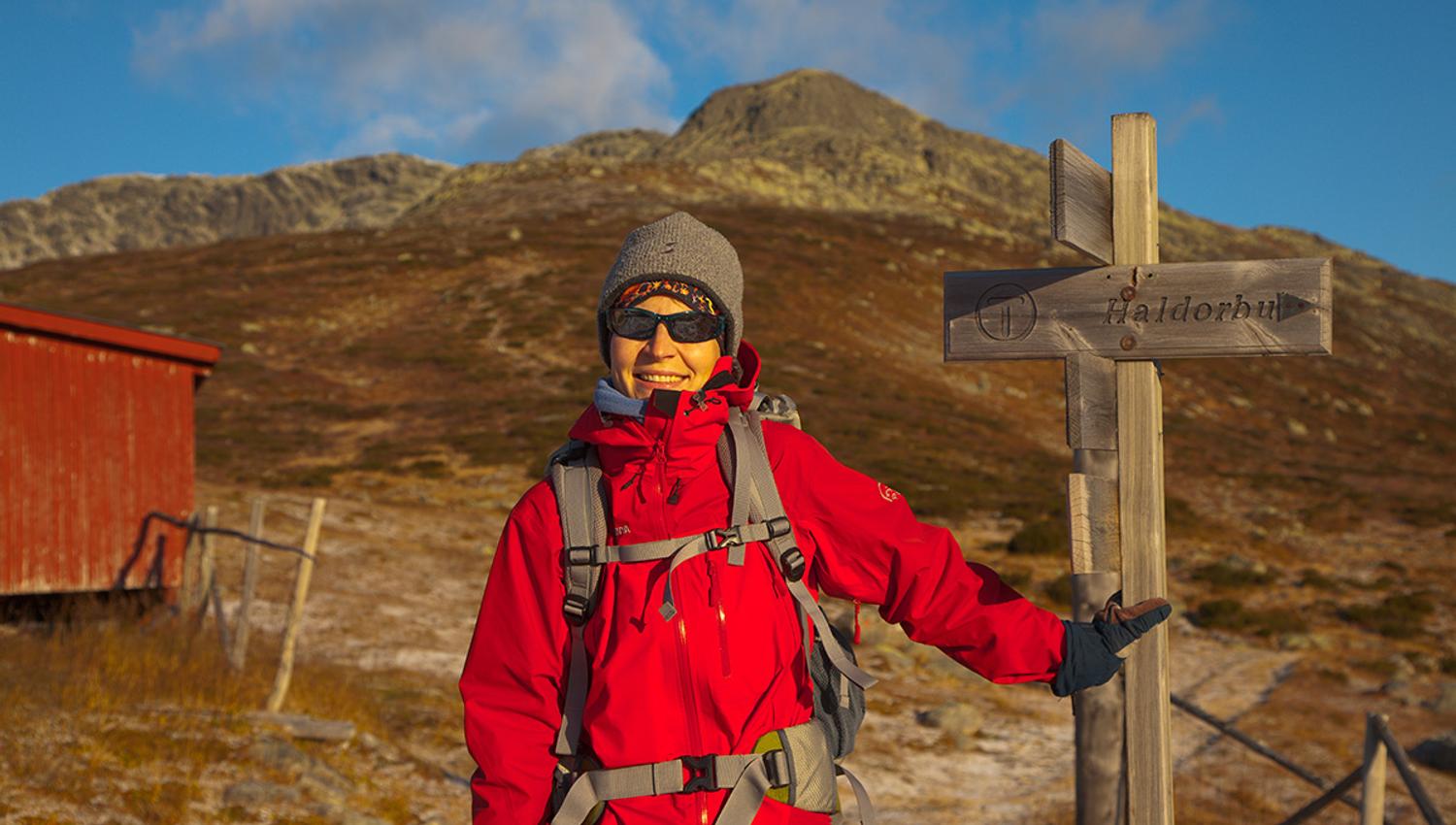Hiker in a red jacket outside the cabin Storeskag at the sign pointing the way towards the summit of Skaget.