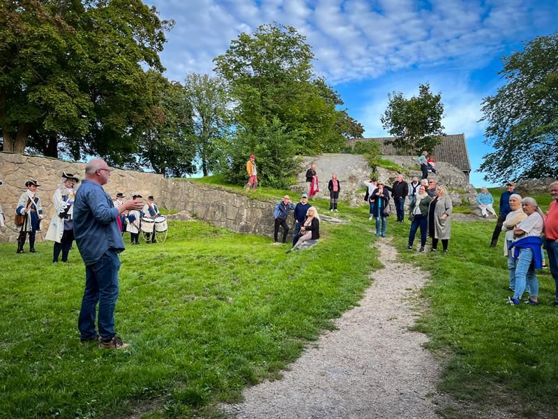 En gruppe mennesker lytter til en formidler på Kongsten Fort, mens Tambouravdelingen i historiske uniformer står i bakgrunnen.