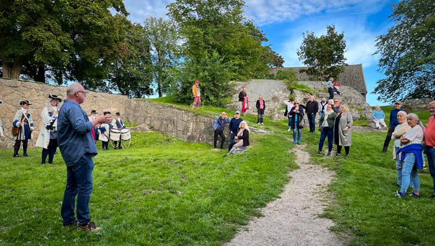 En gruppe mennesker lytter til en formidler på Kongsten Fort, mens Tambouravdelingen i historiske uniformer står i bakgrunnen.