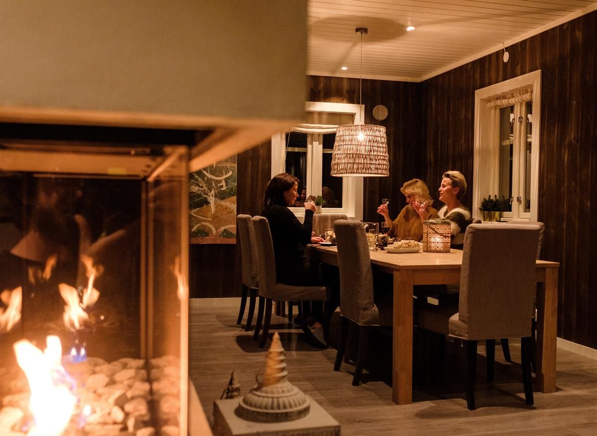 En gruppe mennesker sitter rundt et spisebord innendørs med peis i forgrunnen.