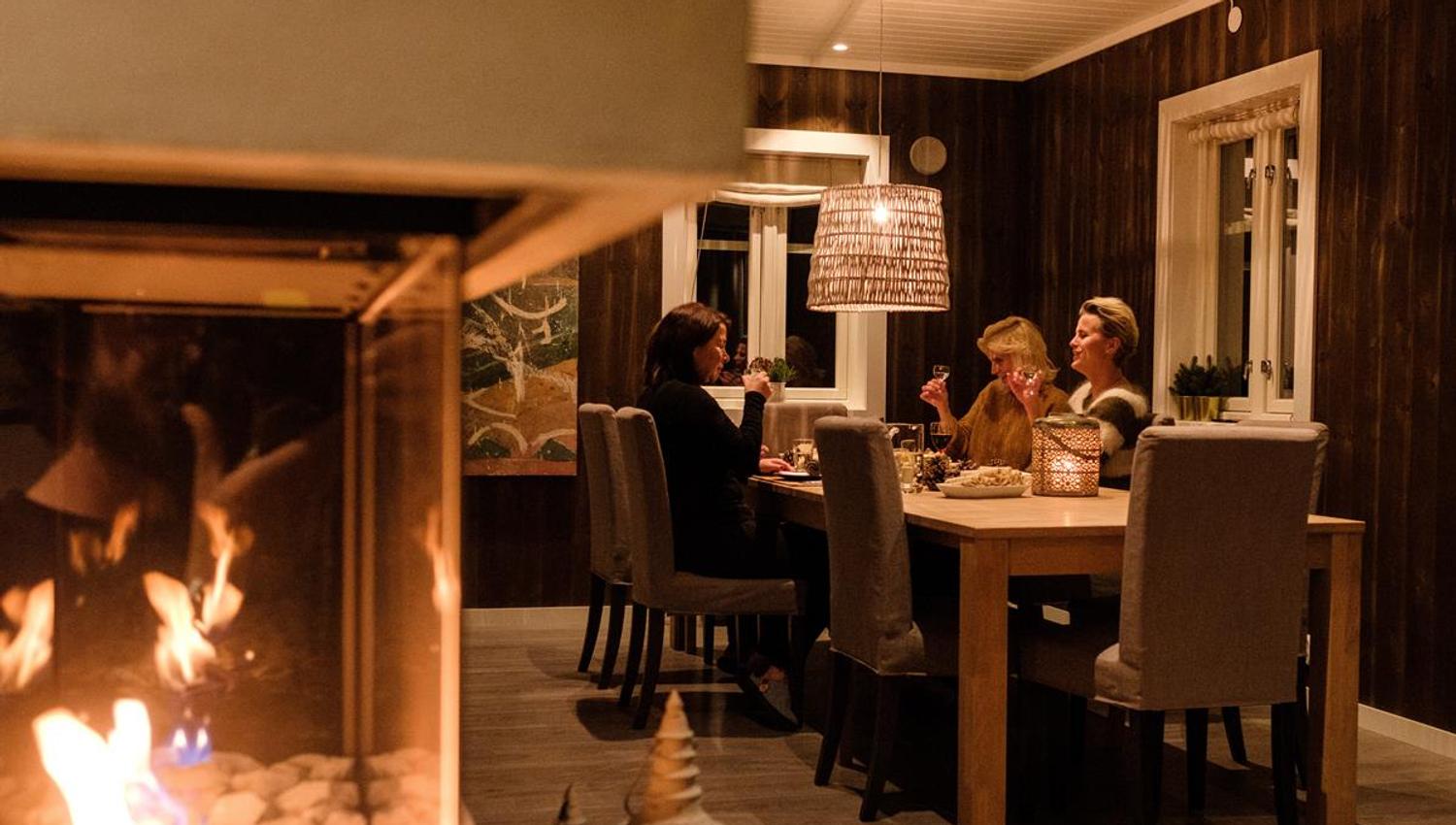 En gruppe mennesker sitter rundt et spisebord innendørs med peis i forgrunnen.