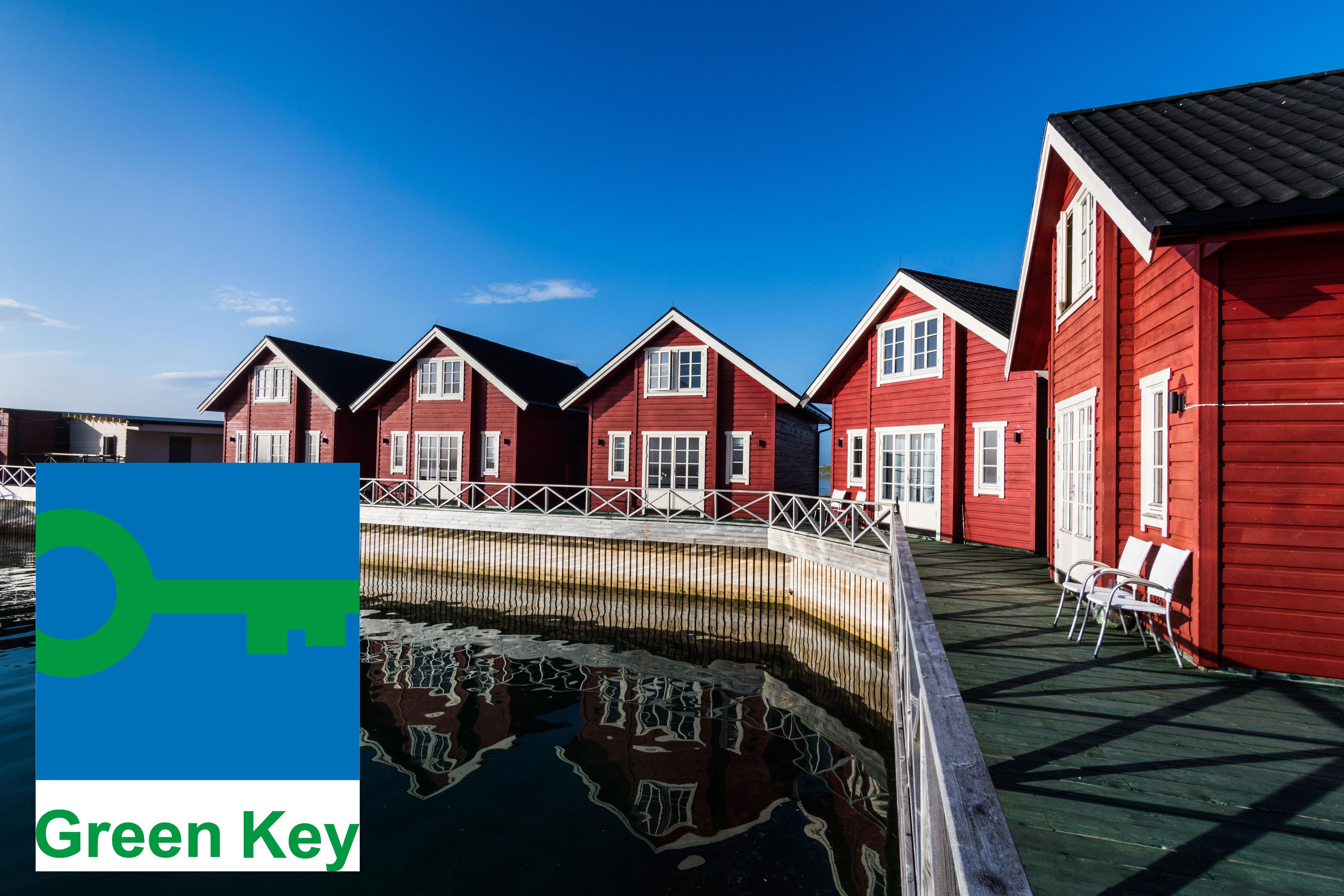 Red wooden houses with white details by the waterfront, reflected in the water. 'Green Key' logo in the bottom left corner