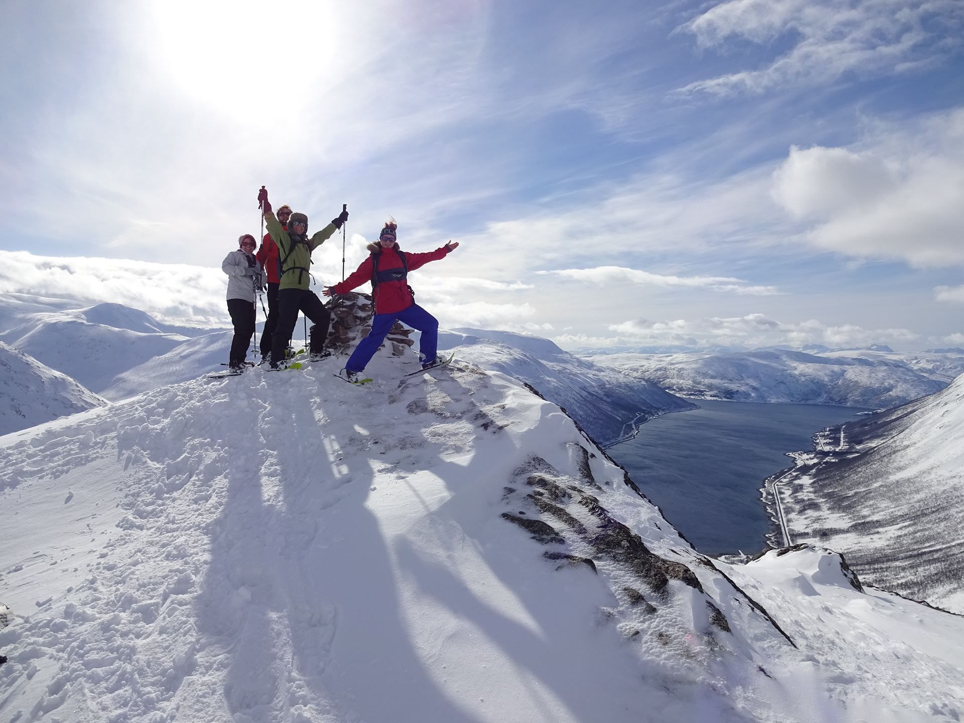 Four people celebrating on a snow-covered mountain peak with a view of a fjord and surrounding mountains.