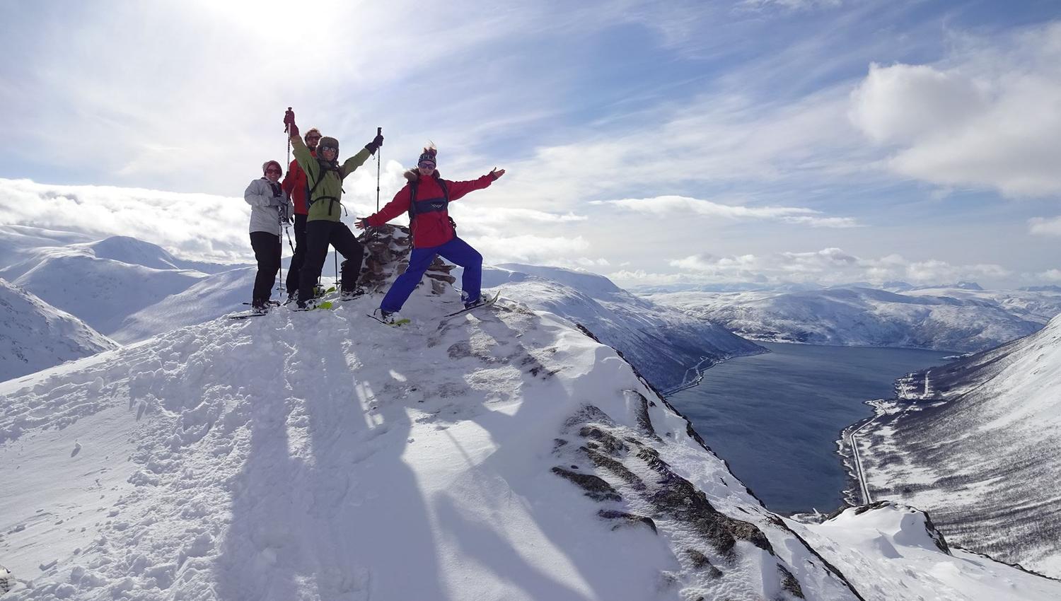 Four people celebrating on a snow-covered mountain peak with a view of a fjord and surrounding mountains.