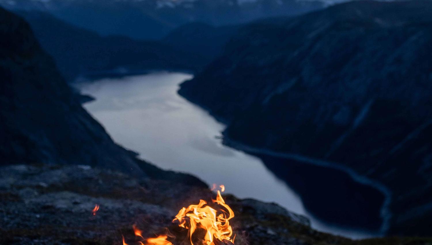 Bålpanne med åpen ild på en fjelltopp ved solnedgang med utsikt over en fjord.