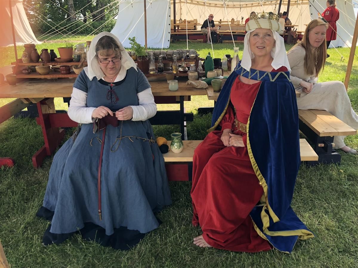 Two people dressed in historical medieval clothing are seated at a wooden table in an outdoor medieval‑themed setting. The table is filled with potter