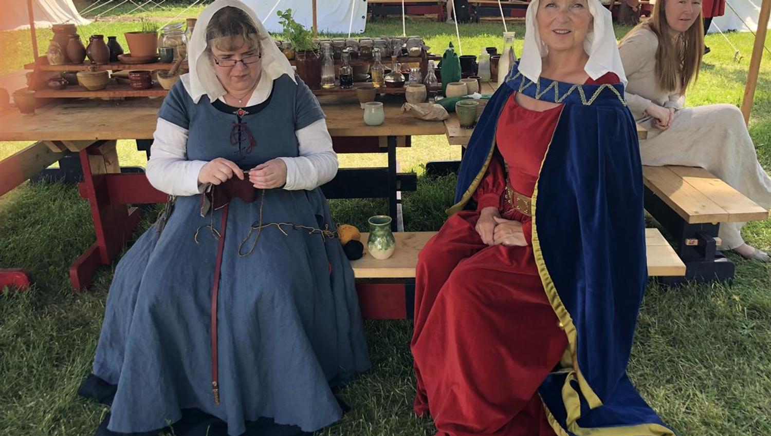 Two people dressed in historical medieval clothing are seated at a wooden table in an outdoor medieval‑themed setting. The table is filled with potter