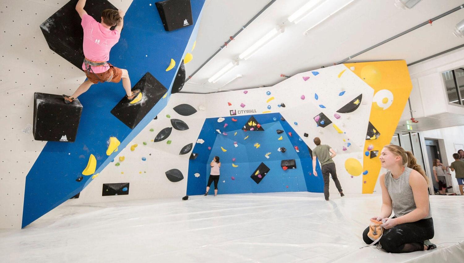 People bouldering in an indoor climbing gym with colourful holds and padded mats.