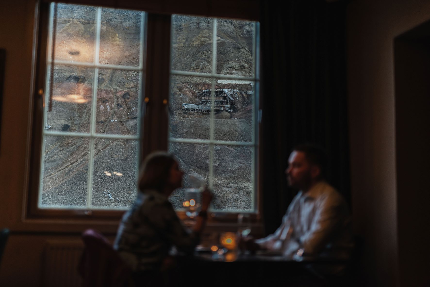 To personer som sitter ved et restaurantbord med utsikt til et fjell og en gammel gruve gjennom et vindu i bakgrunnen