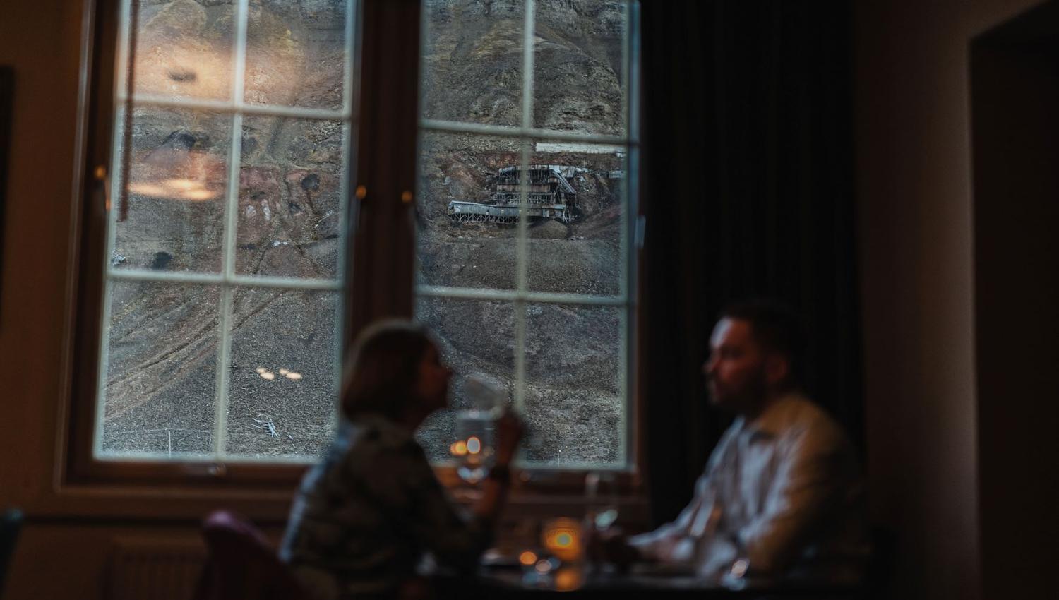 To personer som sitter ved et restaurantbord med utsikt til et fjell og en gammel gruve gjennom et vindu i bakgrunnen