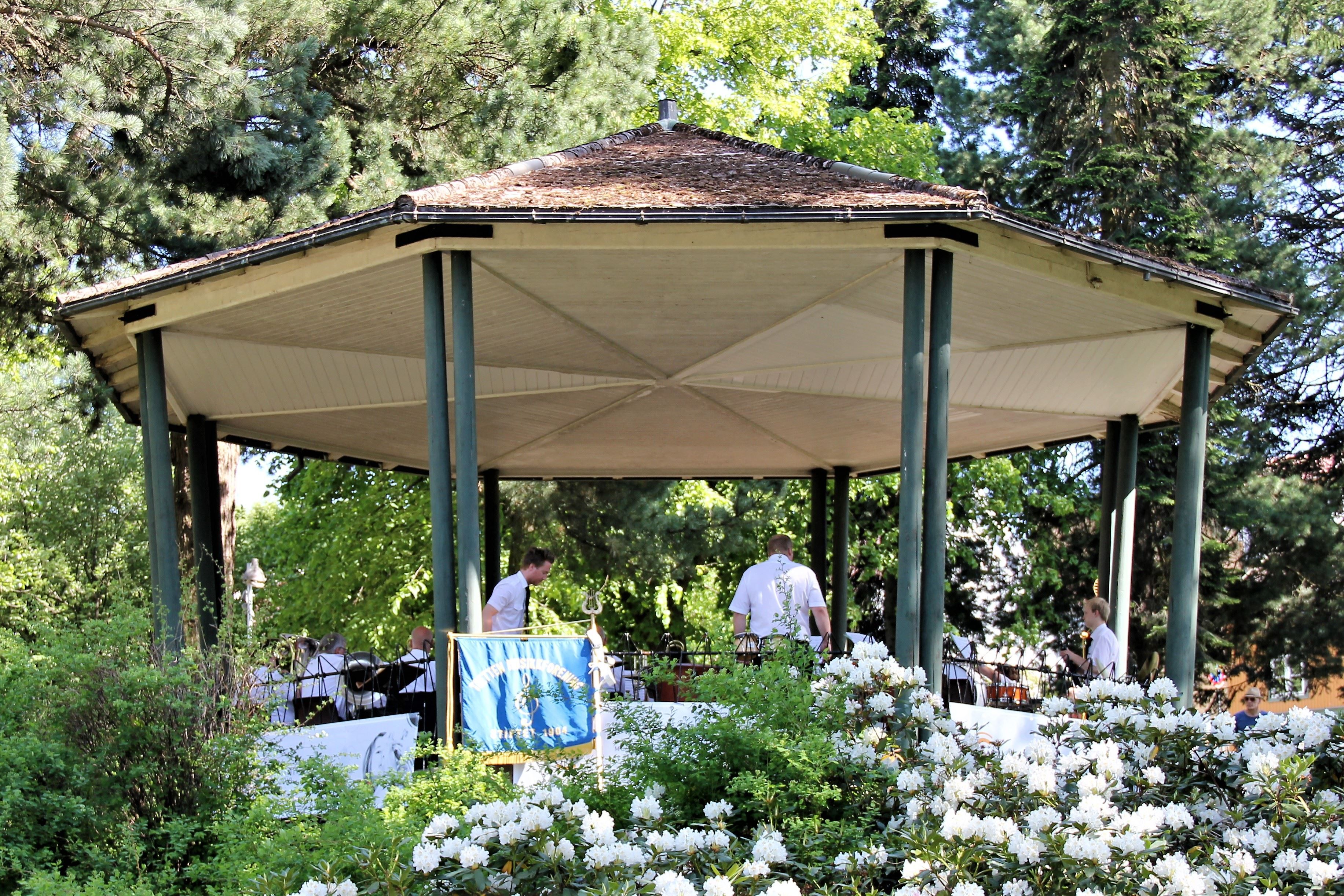 En åttekantet paviljong i en grønn park, med folk i hvite skjorter og blomstrende busker foran..