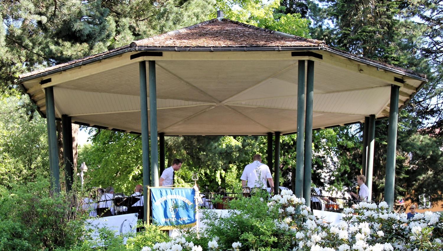 En åttekantet paviljong i en grønn park, med folk i hvite skjorter og blomstrende busker foran..