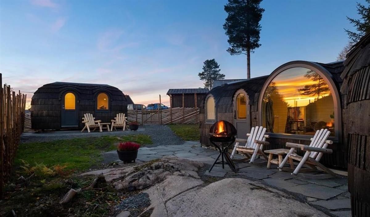 Et uteområde med trehytter, sitteplasser og en tent bålpanne i skumring.