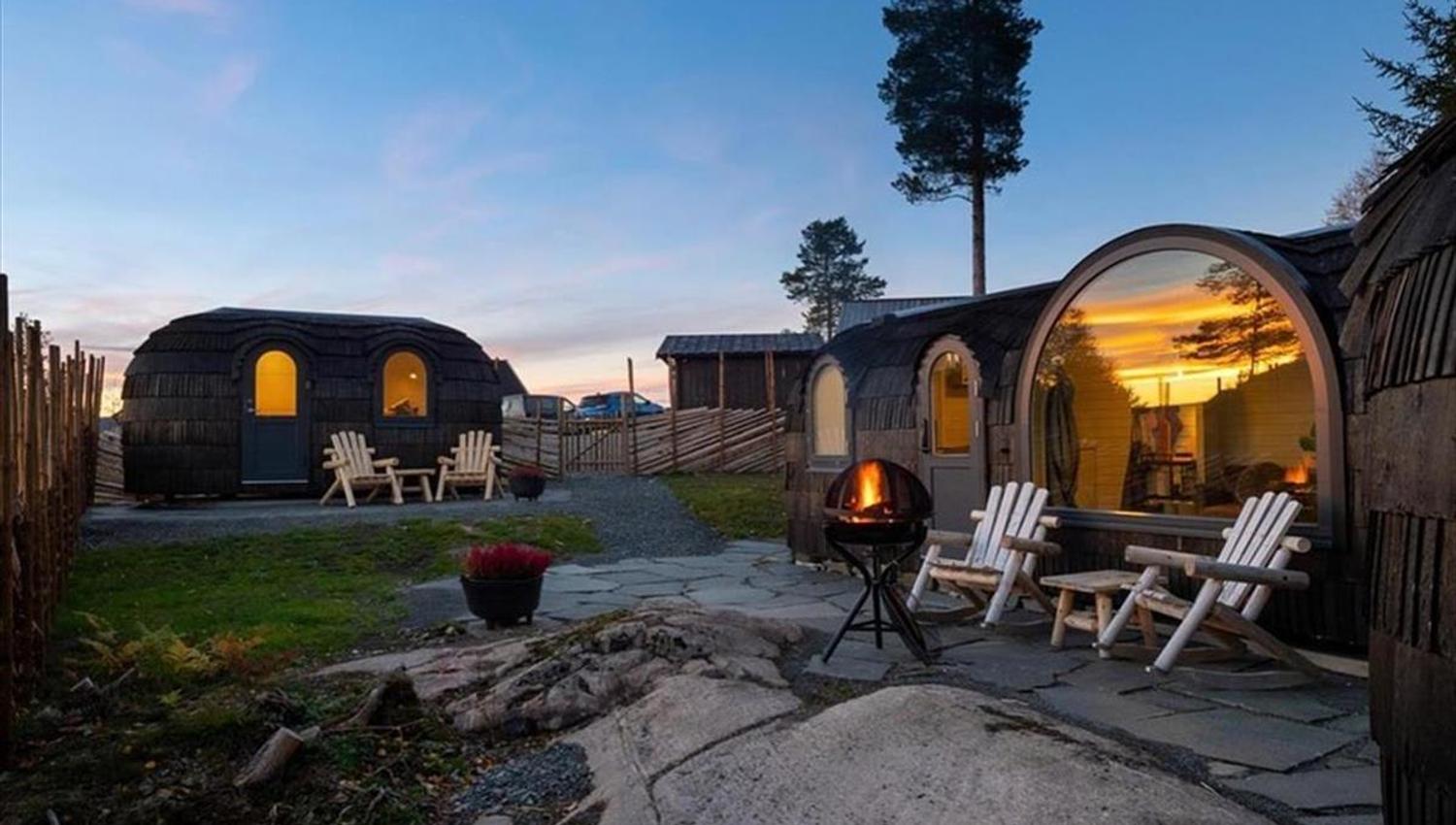 Et uteområde med trehytter, sitteplasser og en tent bålpanne i skumring.