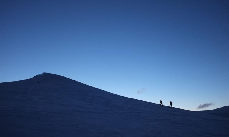 To personer i silhuett som går opp en snødekt fjelltopp, med mørk blå himmel i bakgrunnen