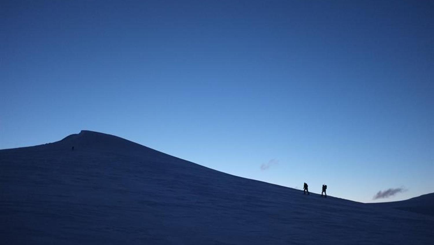 To personer i silhuett som går opp en snødekt fjelltopp, med mørk blå himmel i bakgrunnen