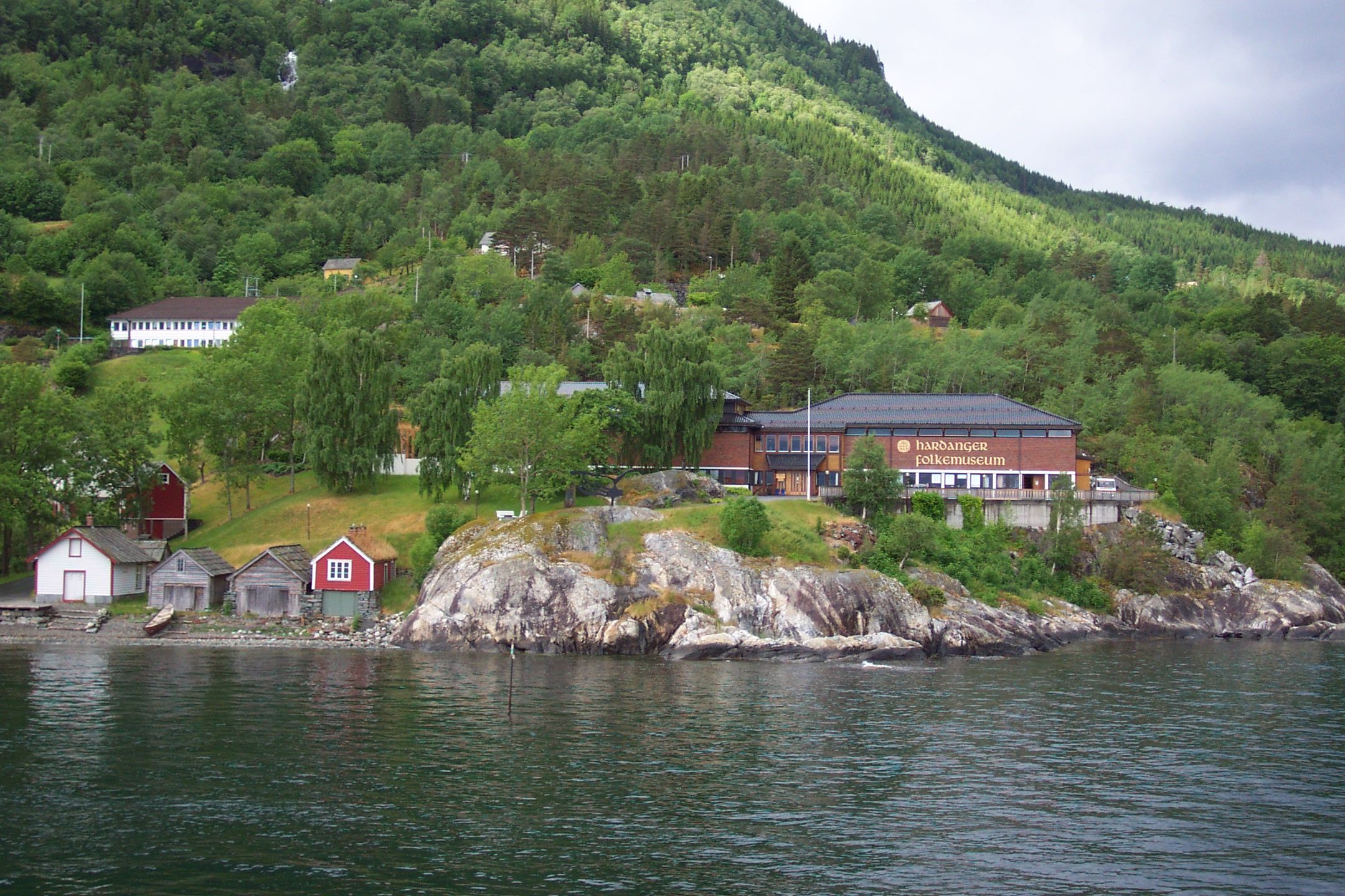 Hardanger folkemuseum sett frå fjorden, omgitt av grøne bakkar og fjell.