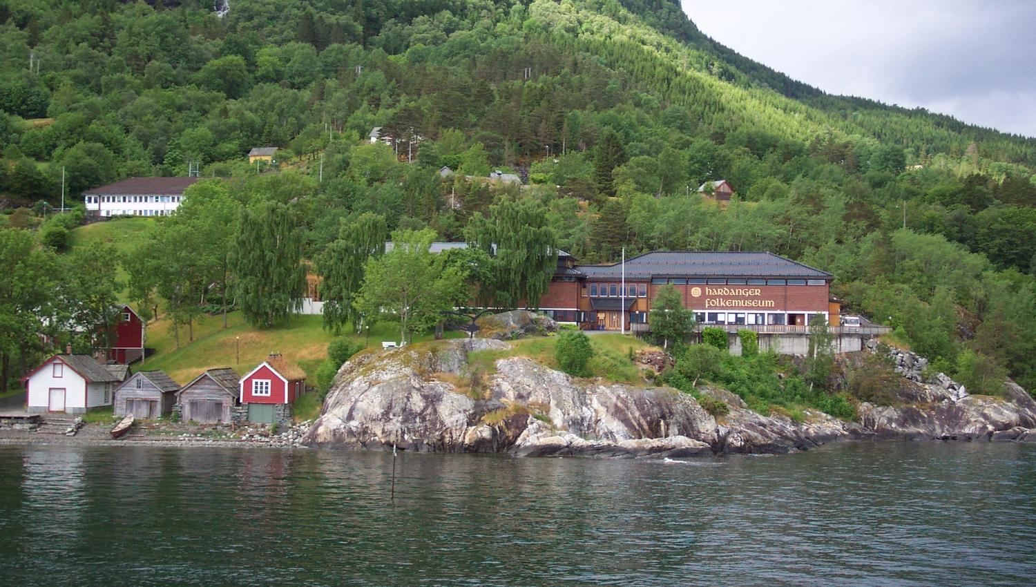 Hardanger folkemuseum sett frå fjorden, omgitt av grøne bakkar og fjell.