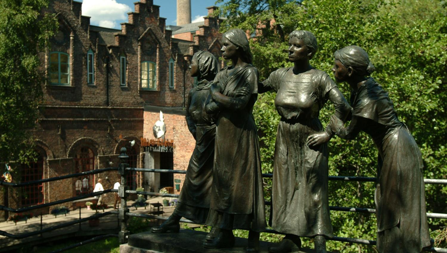 Statue av fire jenter med trær og bygning i bakgrunnen.