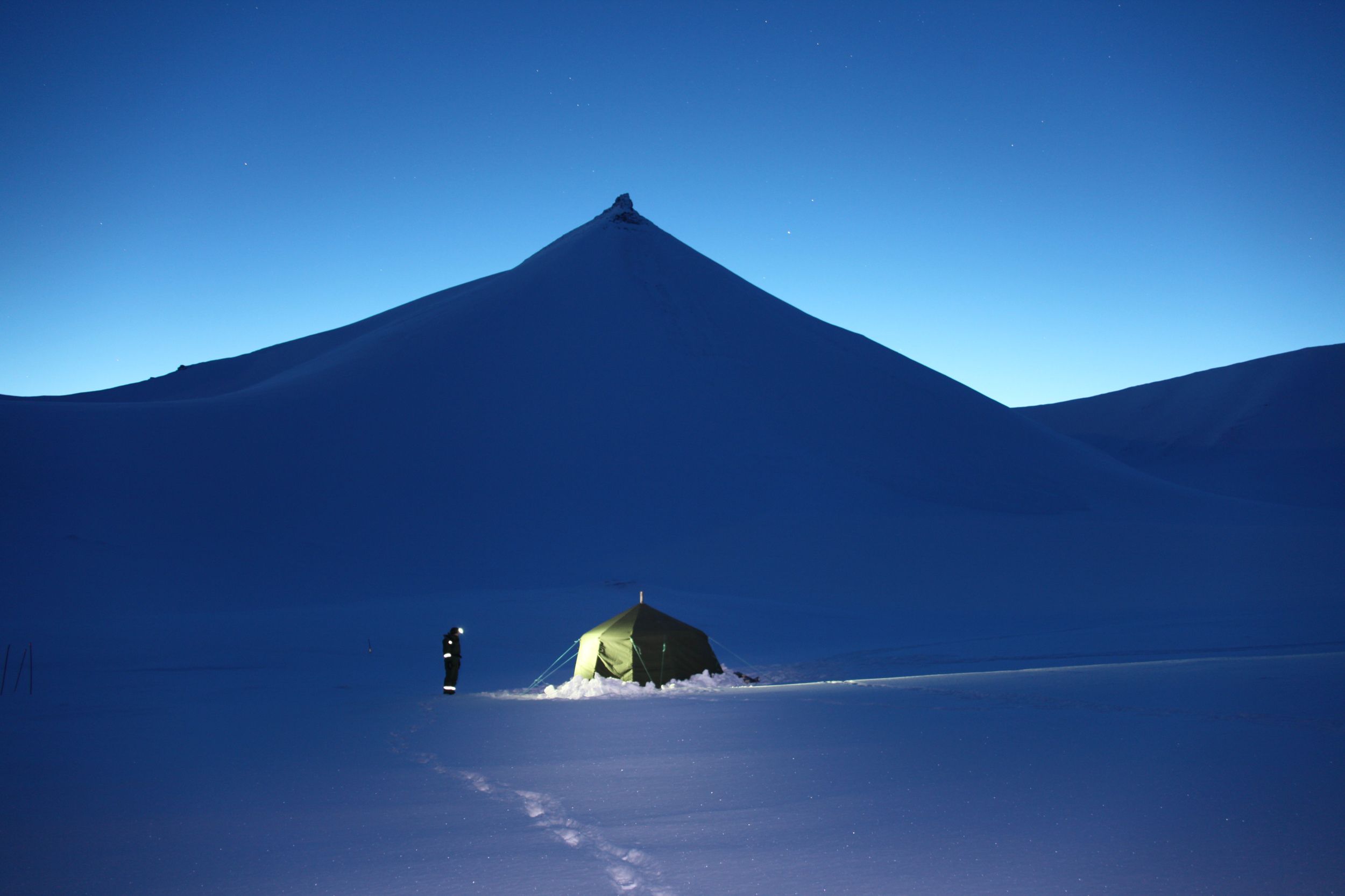 En person med hodelykt utenfor et telt i blåtimen, fjellformasjoner i bakgrunnen.