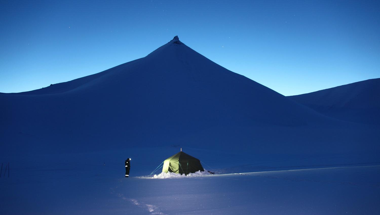 En person med hodelykt utenfor et telt i blåtimen, fjellformasjoner i bakgrunnen.