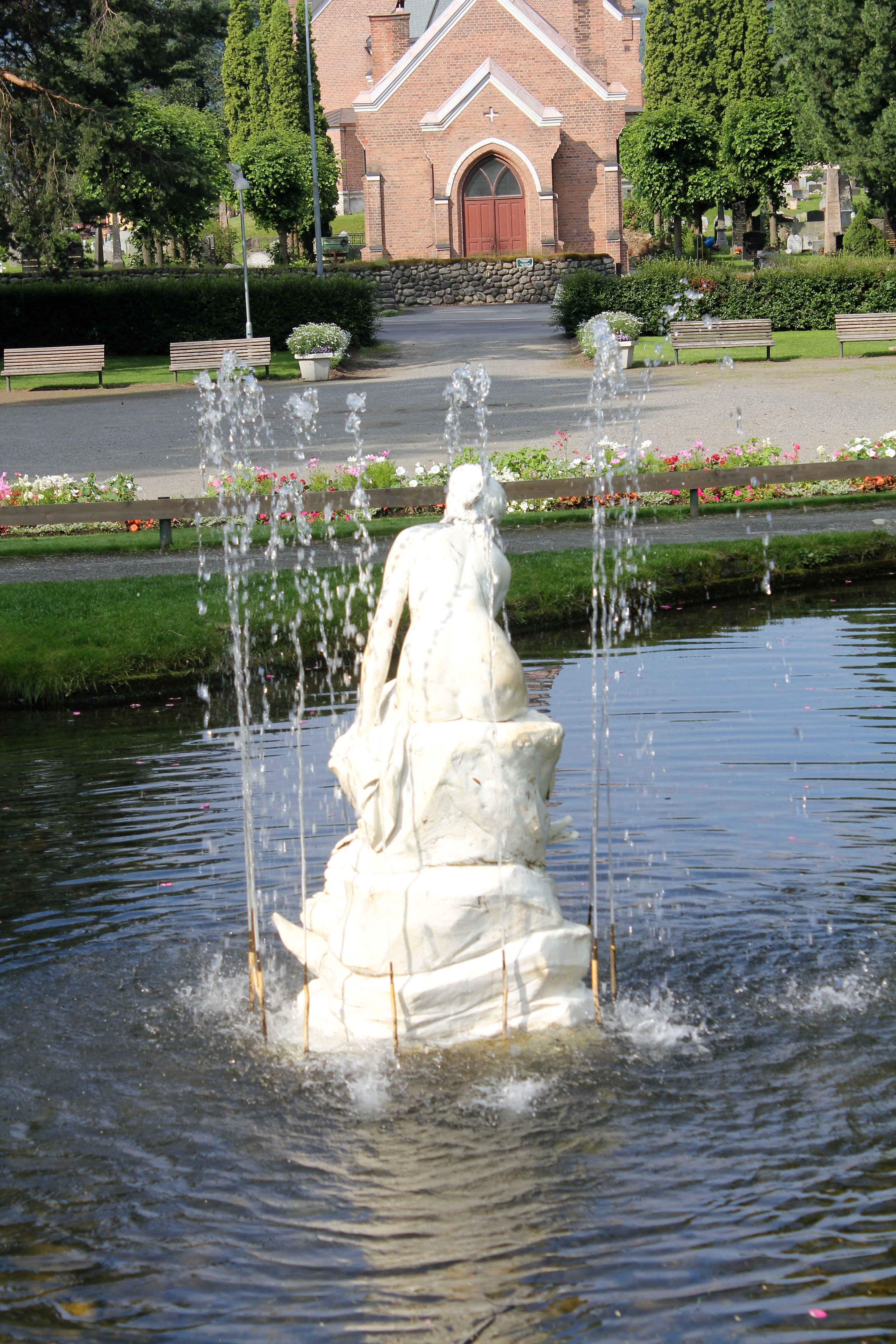 Hvit statue i en fontene med vannsprut i dammen, med blomsterbed og en mursteinskirke i bakgrunnen..