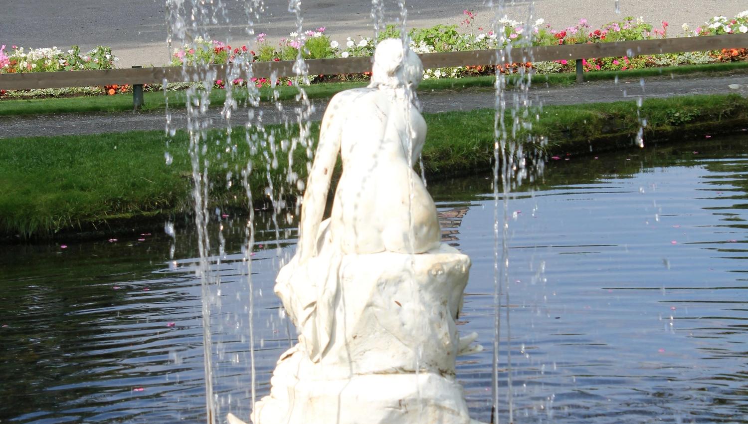 Hvit statue i en fontene med vannsprut i dammen, med blomsterbed og en mursteinskirke i bakgrunnen..