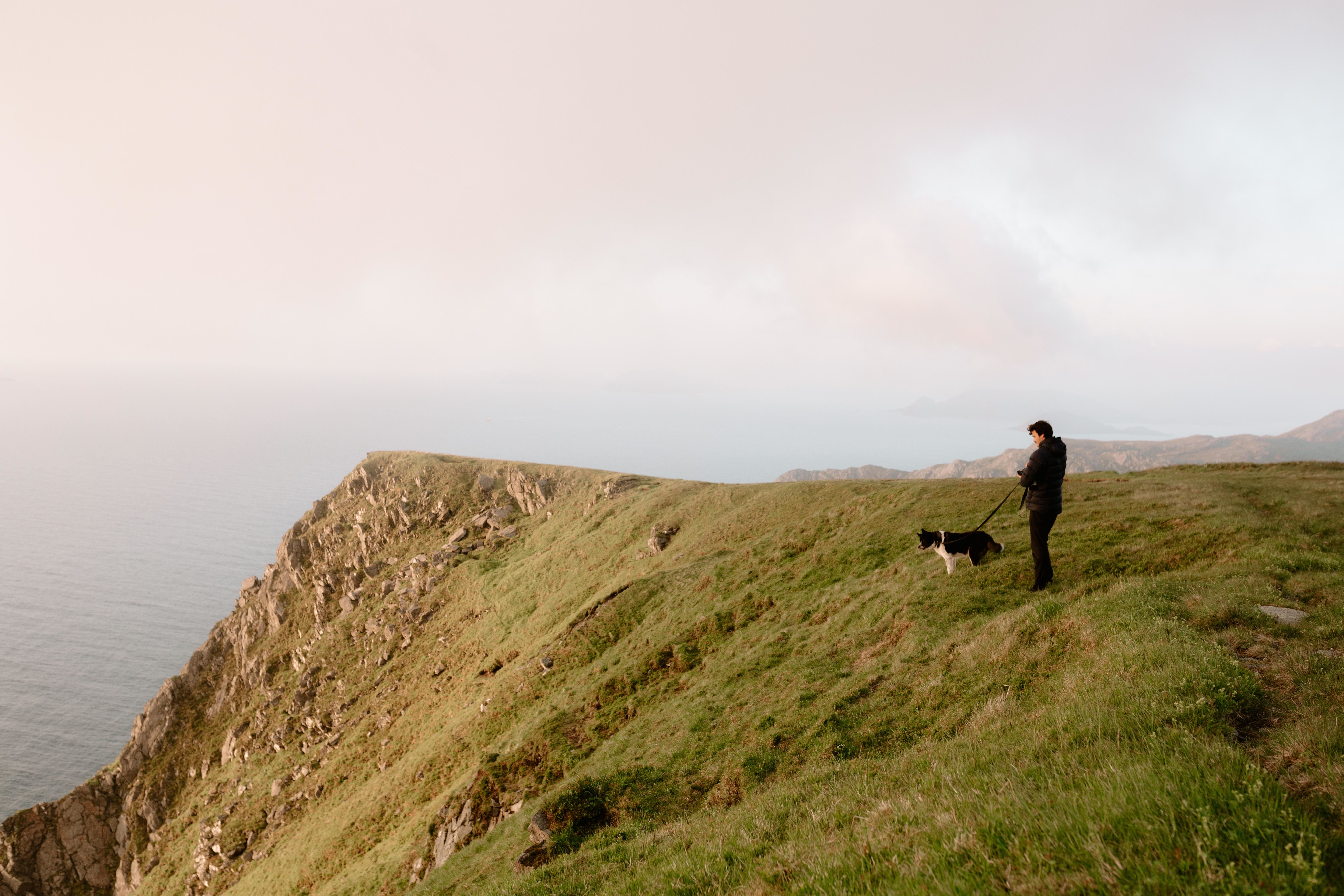 Ein person i mørk jakke står på ei grøn klippe ved havet med ein svartkvit hund i band under disig himmel..