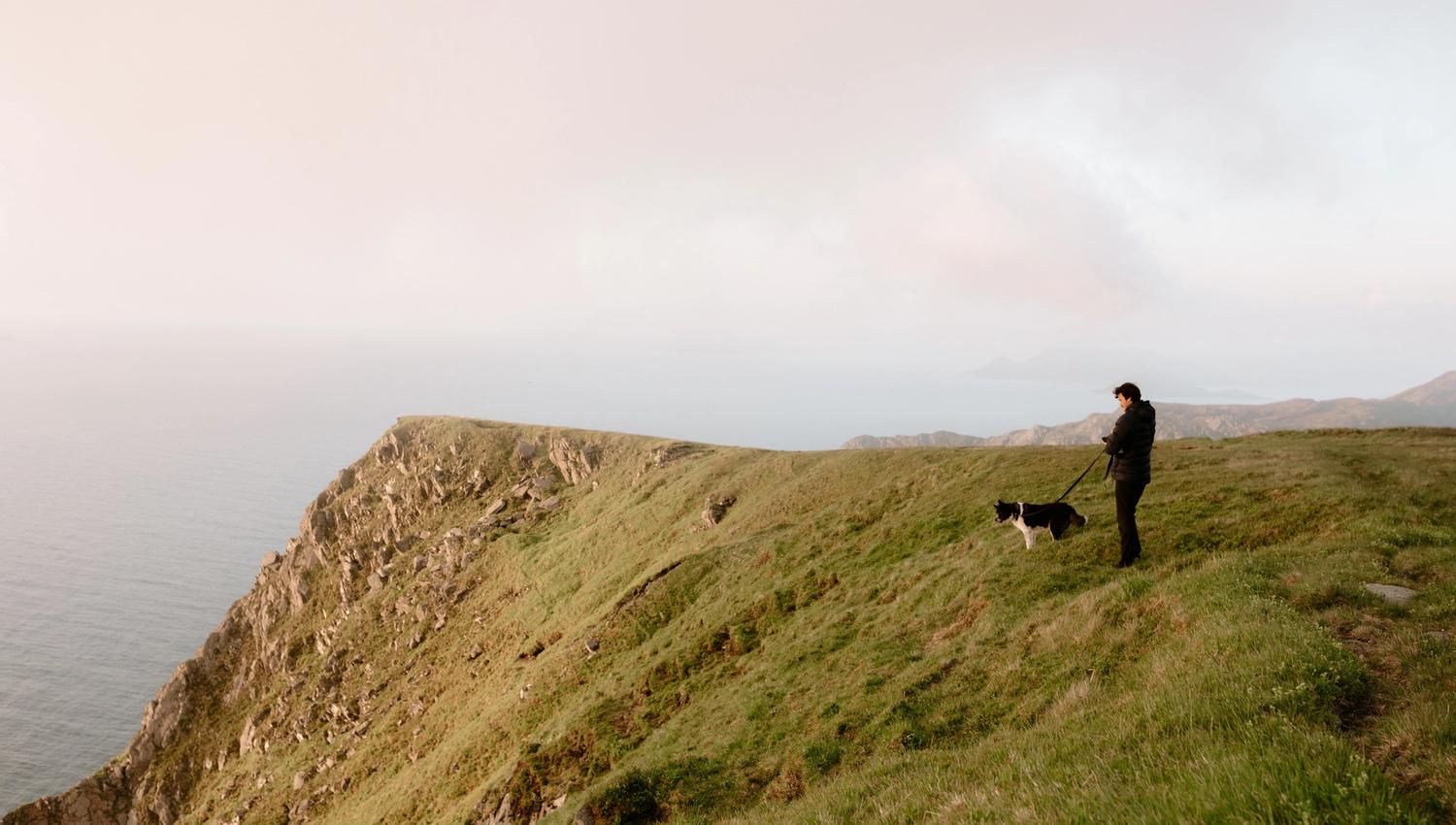 Ein person i mørk jakke står på ei grøn klippe ved havet med ein svartkvit hund i band under disig himmel..