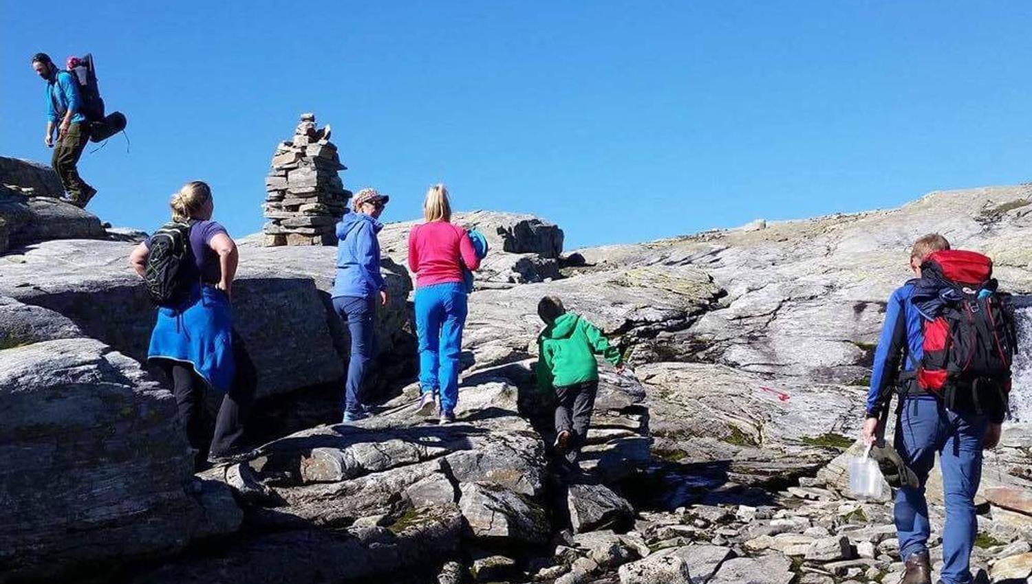 Familie på tur langs steinete fjellterreng ved Hallet grøne i Hardanger, med klar blå himmel og vardar som viser vegen.