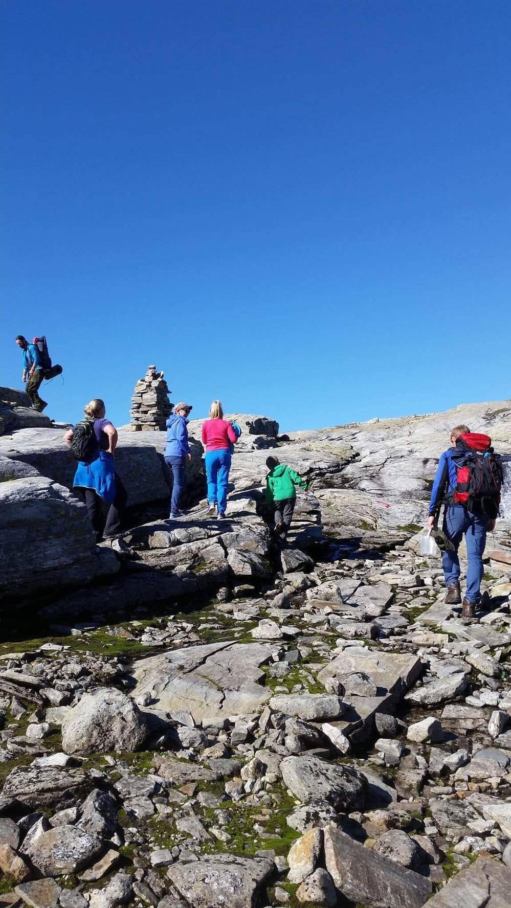 Family hiking across rocky mountain terrain at Hallet Grøne in Hardanger, Norway, with cairns marking the trail under a clear blue sky.