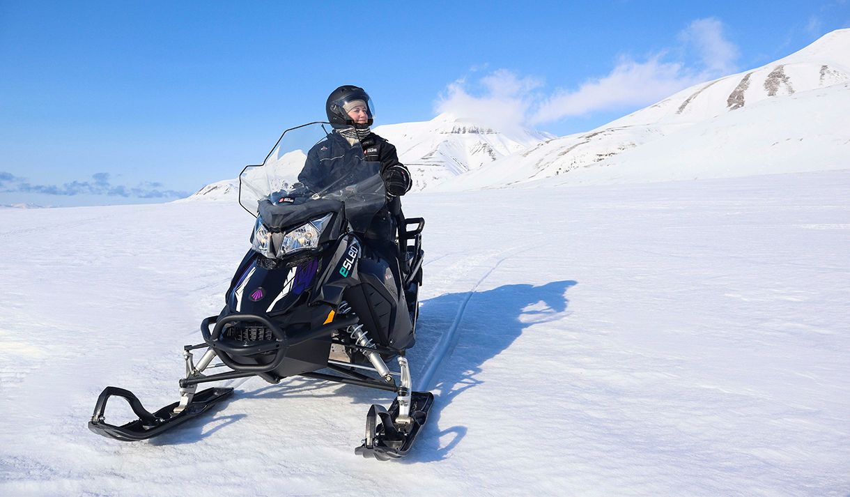 En smilende person i snøscooterutstyr som kjører en elektrisk snøscooter gjennom et snødekt landskap