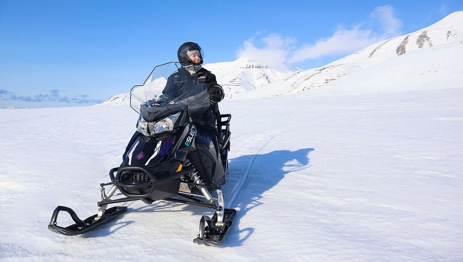 En smilende person i snøscooterutstyr som kjører en elektrisk snøscooter gjennom et snødekt landskap