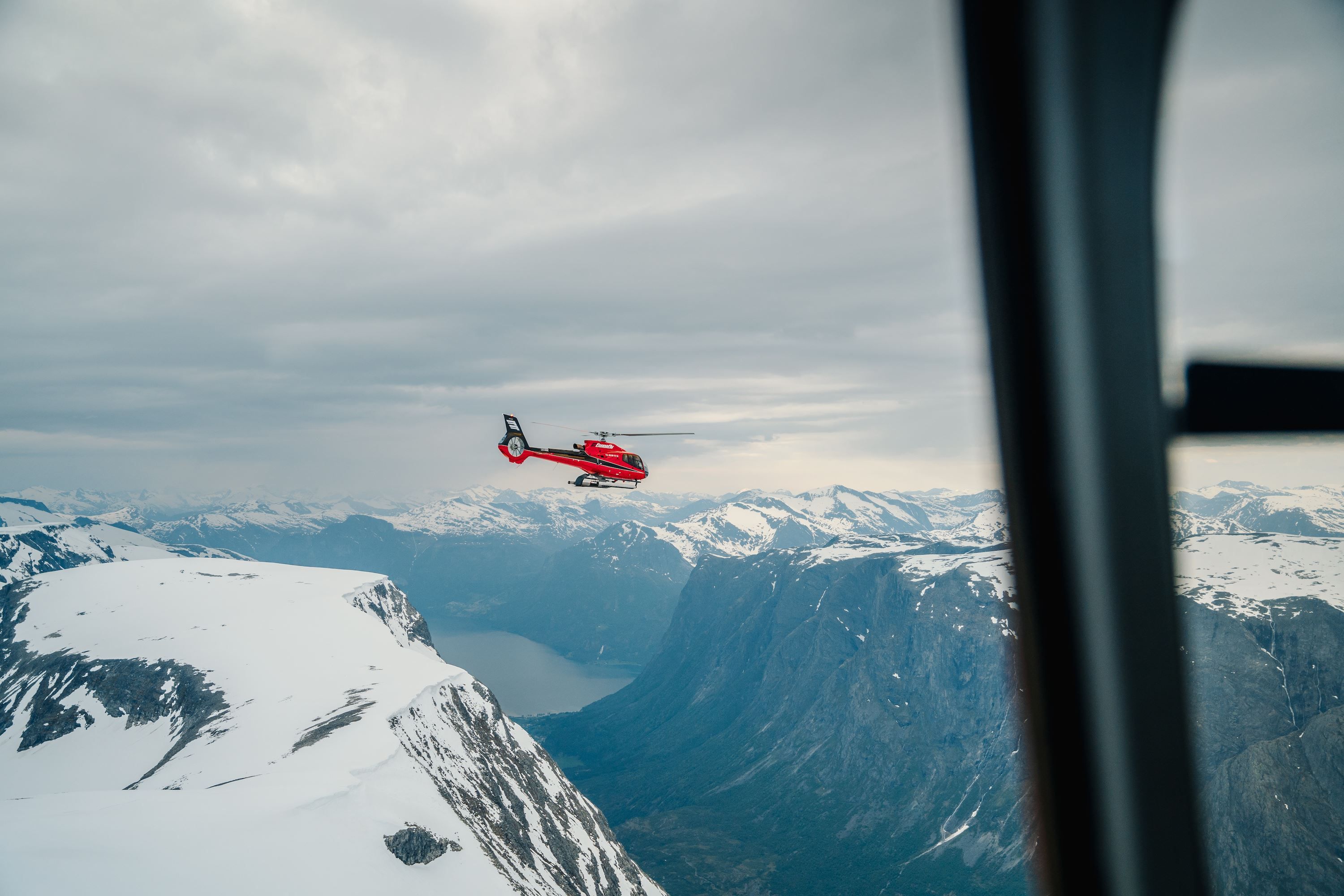 Fjord Helikopter  -Eit raud helikopter flyg over snødekte fjell og ein fjord under ein overskya himmel, sett frå eit vindauge..