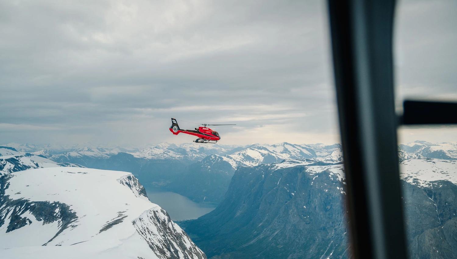 Fjord Helikopter -Eit raud helikopter flyg over snødekte fjell og ein fjord under ein overskya himmel, sett frå eit vindauge..