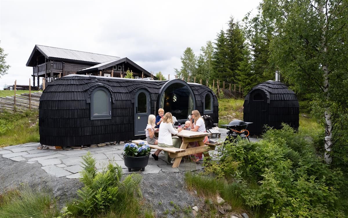 Family gathered at a picnic table in front of wooden cabin pods in a green forest setting.