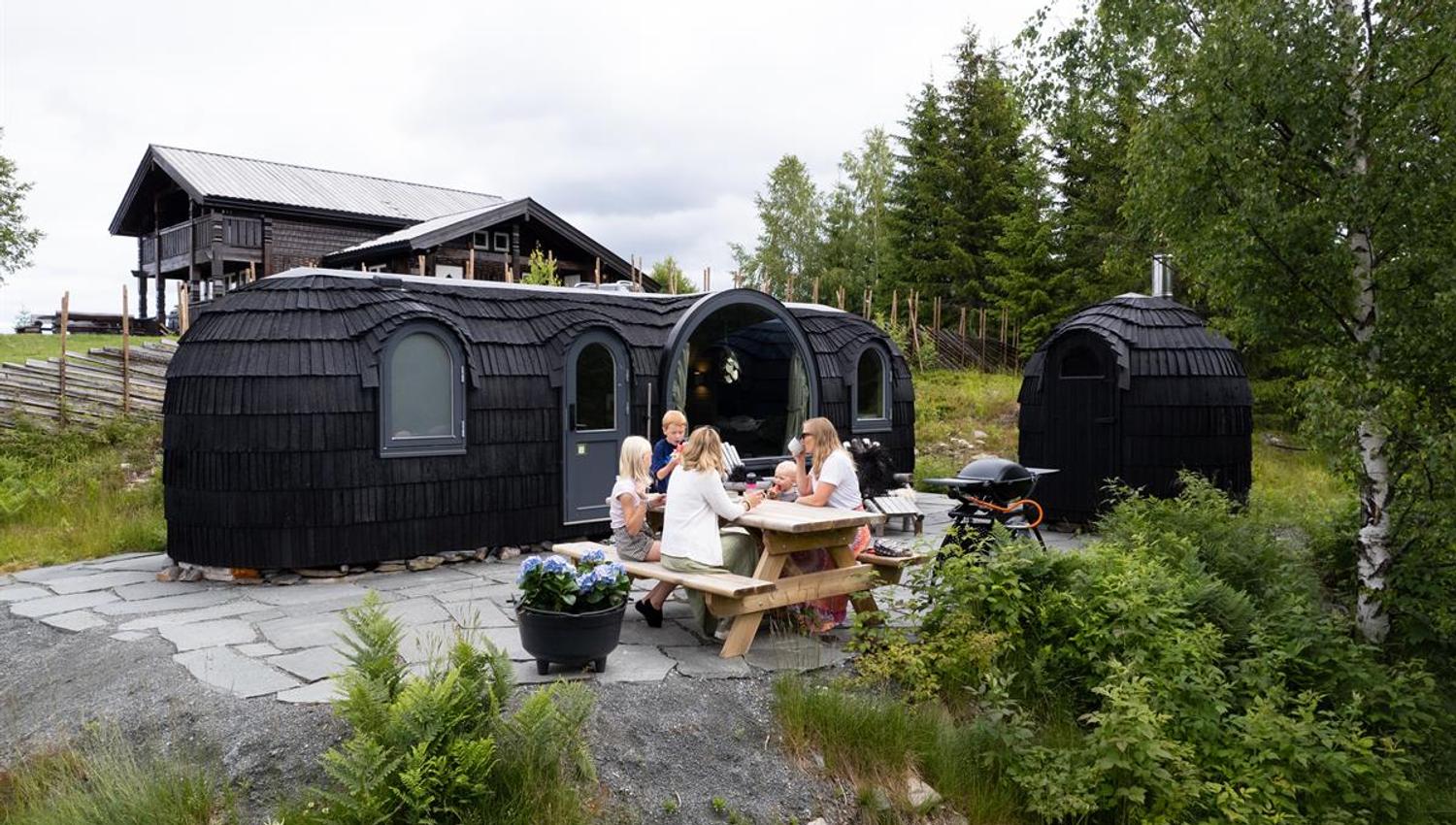 Family gathered at a picnic table in front of wooden cabin pods in a green forest setting.